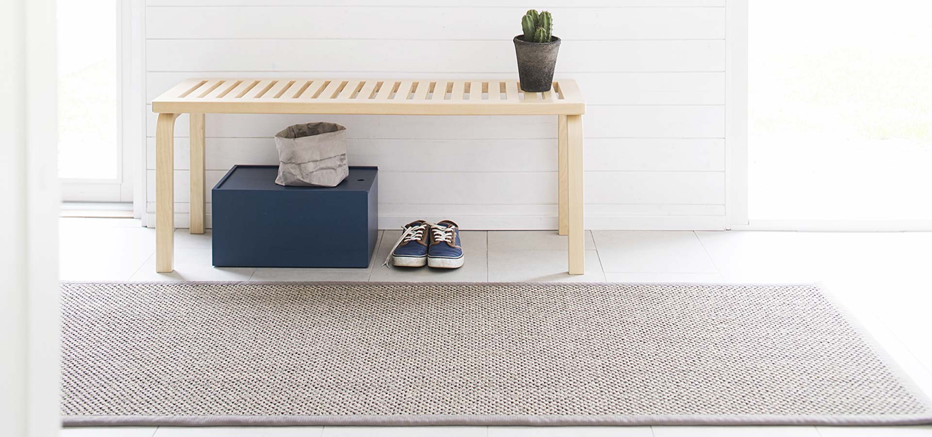 Vaaleanharmaa matto eteisess&auml;, jossa on muutama pari kenki&auml; ja puinen penkki.